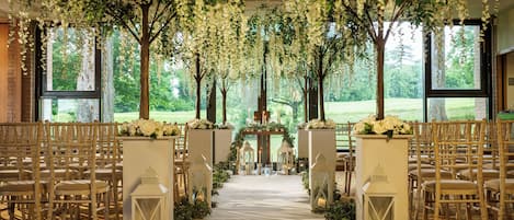An indoor venue with a floral canopy, rows of chairs, and a central aisle leading to a stage.