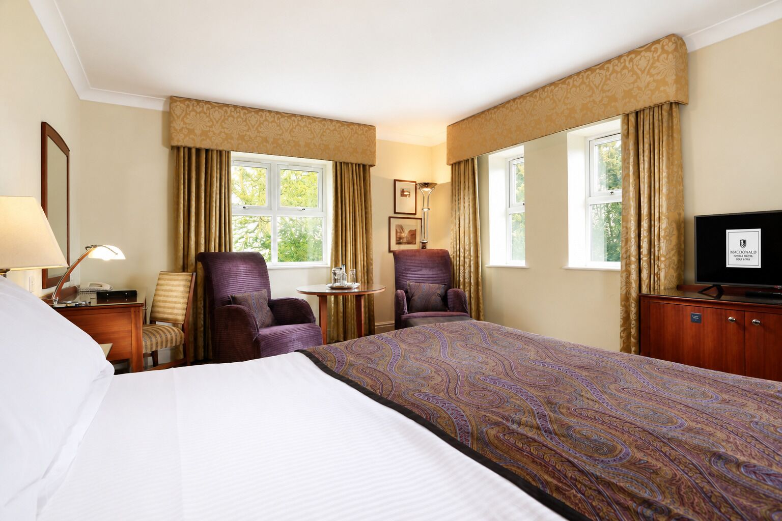 Een hotelkamer met een groot bed, twee fauteuils, een bureau met lamp, een televisie en twee ramen voorzien van gordijnen.