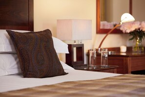 Een netjes opgemaakt bed met een gestreept kussen, een bedlamp en een vaas met bloemen op een nachtkastje van hout.