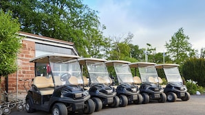 A row of golf carts parked in a lot with a brick building and trees in the background.