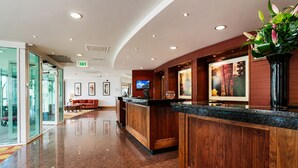 A hotel reception area with a wooden reception desk, a television, framed artwork on the walls, and a glass door entrance.