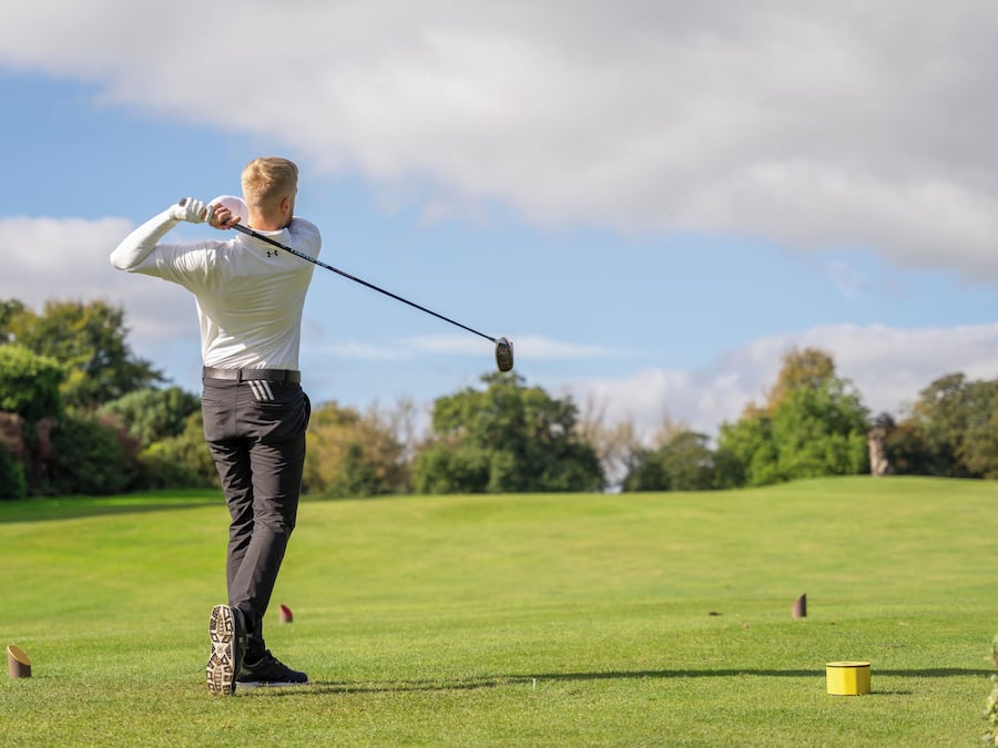 Un golfista en pleno swing en un campo de golf, con árboles y un cielo despejado al fondo.