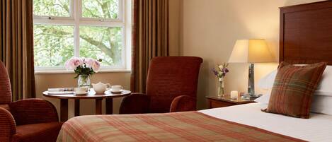 Ein Hotelzimmer mit einem Bett, zwei Sesseln, einem kleinen Tisch mit Blumenvase, einer Lampe und einem Fenster mit Vorhängen.