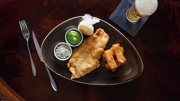 A plate of fish and chips with two dipping sauces, a glass of beer, and a fork and knife on a wooden table.