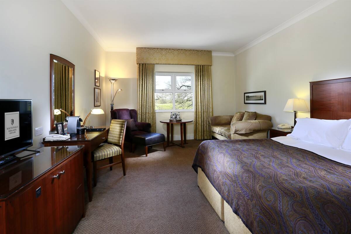 A hotel room with a bed, a desk with a television, a chair, a small table, and a window with curtains.