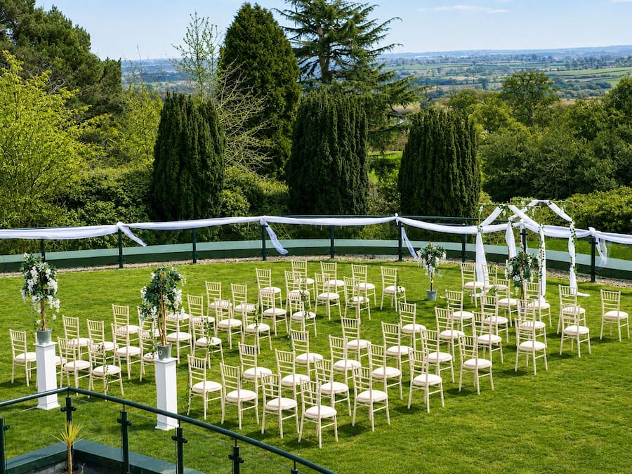 An outdoor wedding venue with rows of white chairs, a white archway, and a green lawn.