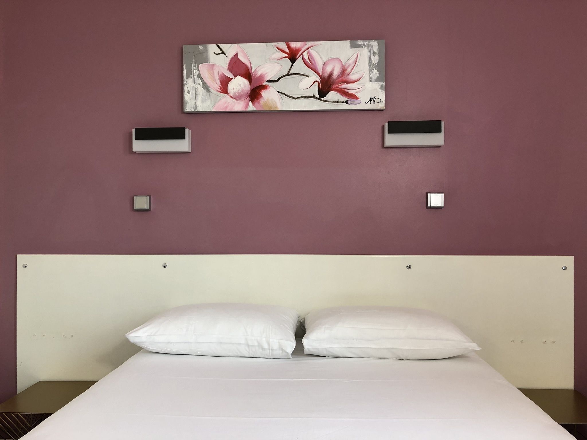 A bedroom with a white bed, two bedside tables, a wall-mounted artwork of pink flowers, and two wall-mounted light switches.
