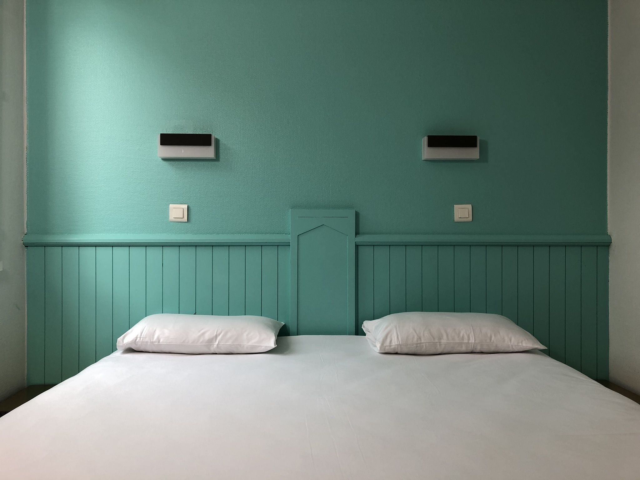 Habitación con paredes color turquesa, cabecera de madera, dos almohadas blancas y dos interruptores de luz fijos a la pared.