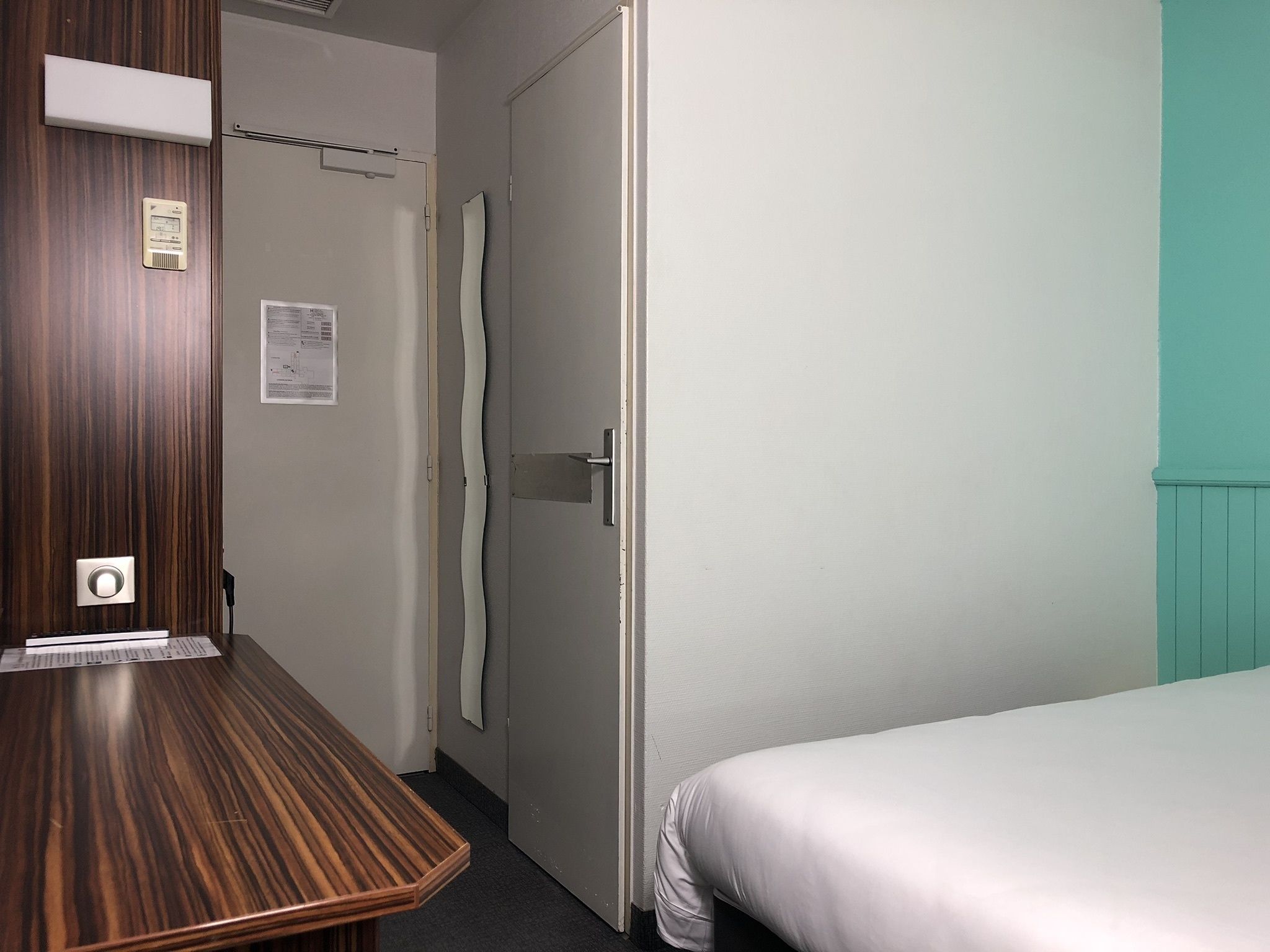 A hotel room with a wooden desk, a bed, a door, and a wall-mounted thermostat.
