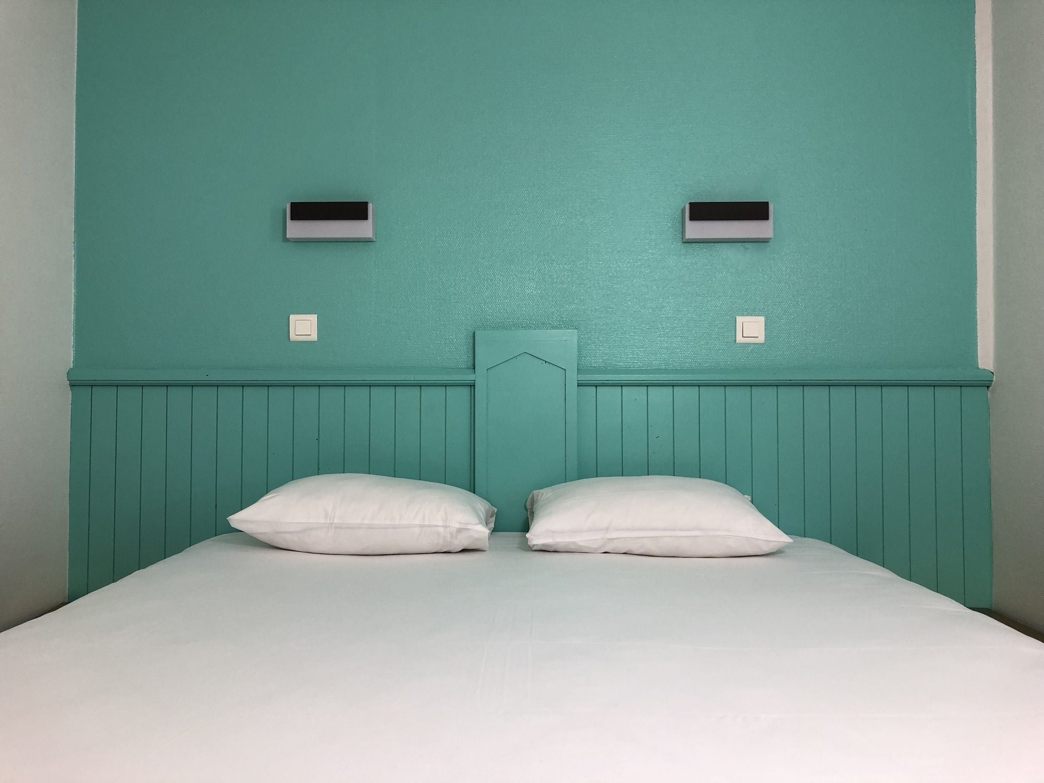 A room with teal walls, a wooden headboard, two white pillows, and two wall-mounted light switches.