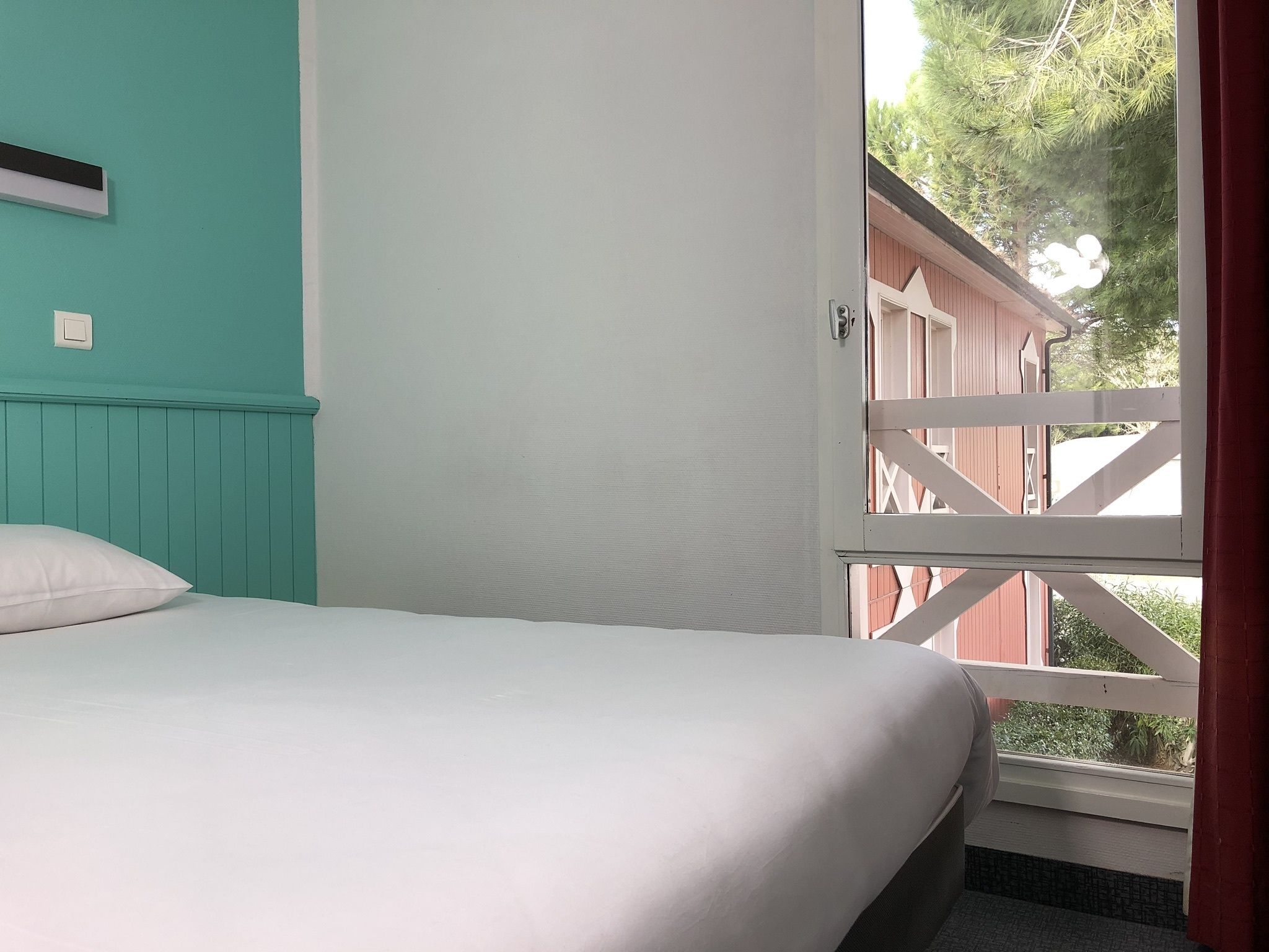 A room with a bed, a window with a view of a wooden deck and trees, and a light switch on the wall.