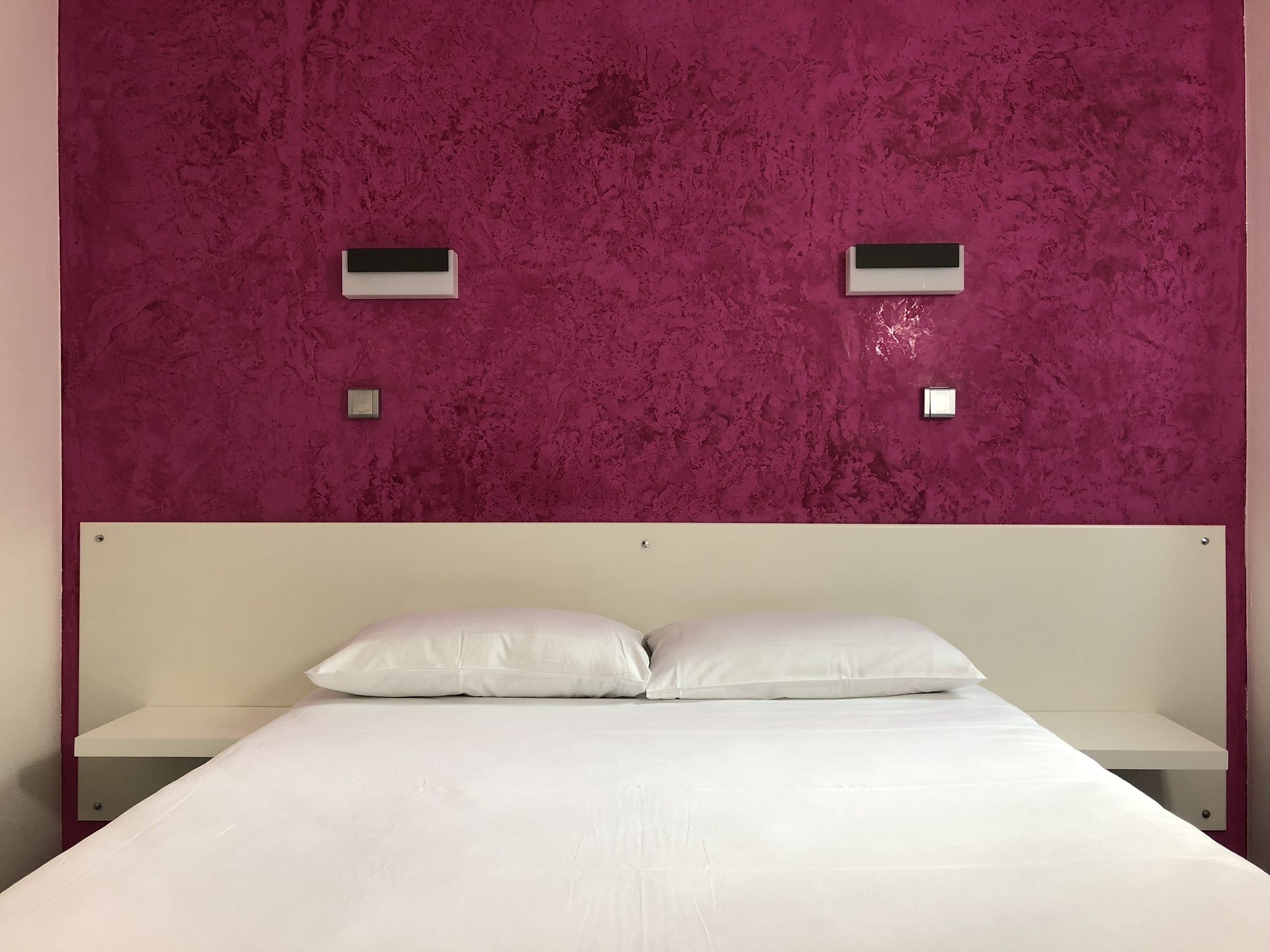 Una habitación con cama, ropa de cama blanca y pared rosa con textura.
