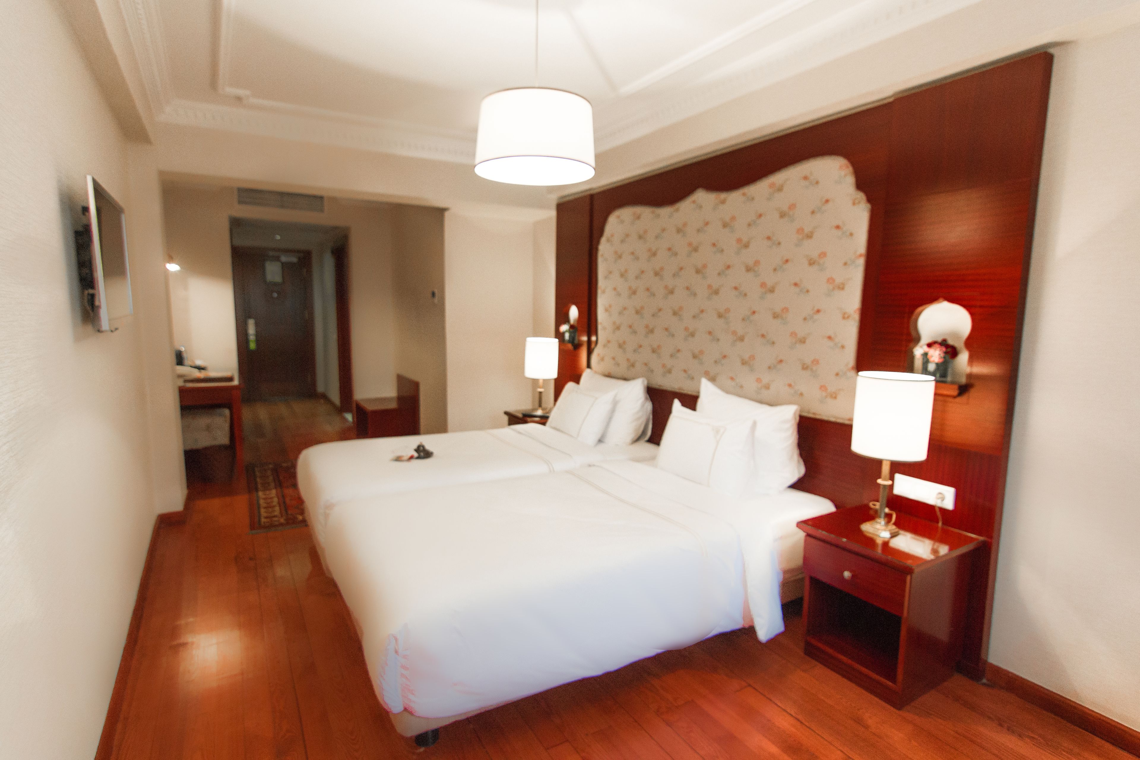 Habitación de hotel con una cama grande, suelo de madera, cabecera con estampado floral, lámparas de noche y una mesita con lámpara.