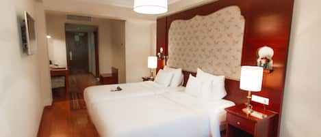 Habitación de hotel con una cama grande, suelo de madera, cabecera con estampado floral, lámparas de noche y una mesita con lámpara.