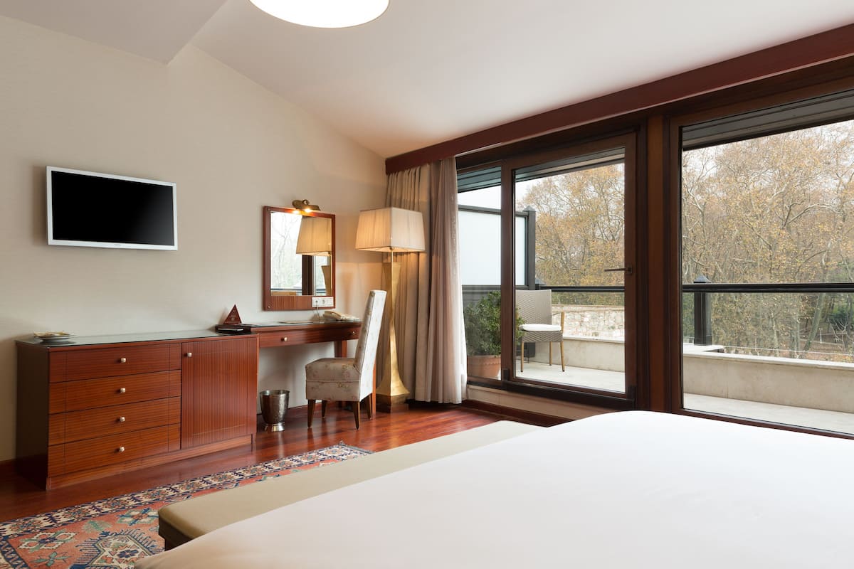 A hotel room with a bed, a desk with a chair, a television, a large window, and a view of trees.