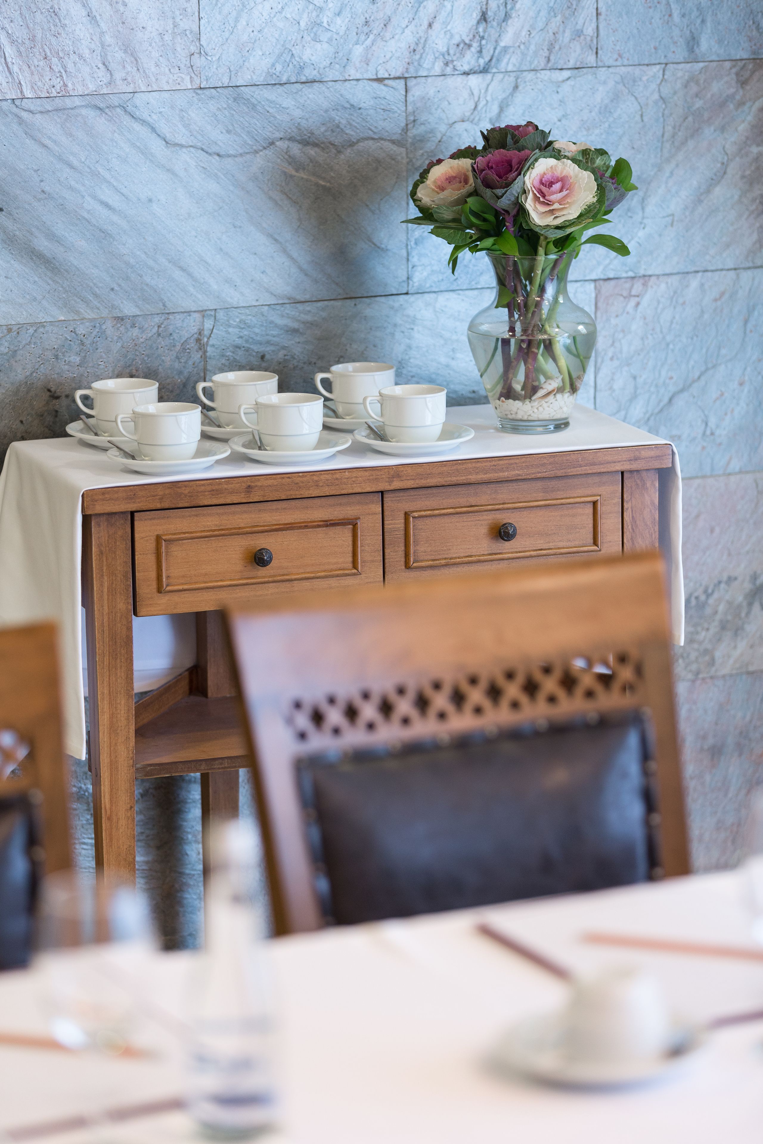 A dining table with a white tablecloth, a vase with flowers, and several white cups and saucers.