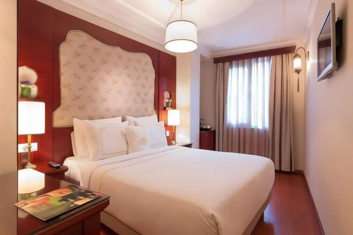 A hotel room with a large bed, two bedside tables, a nightstand, a flat-screen TV, a window with curtains, and a chandelier.