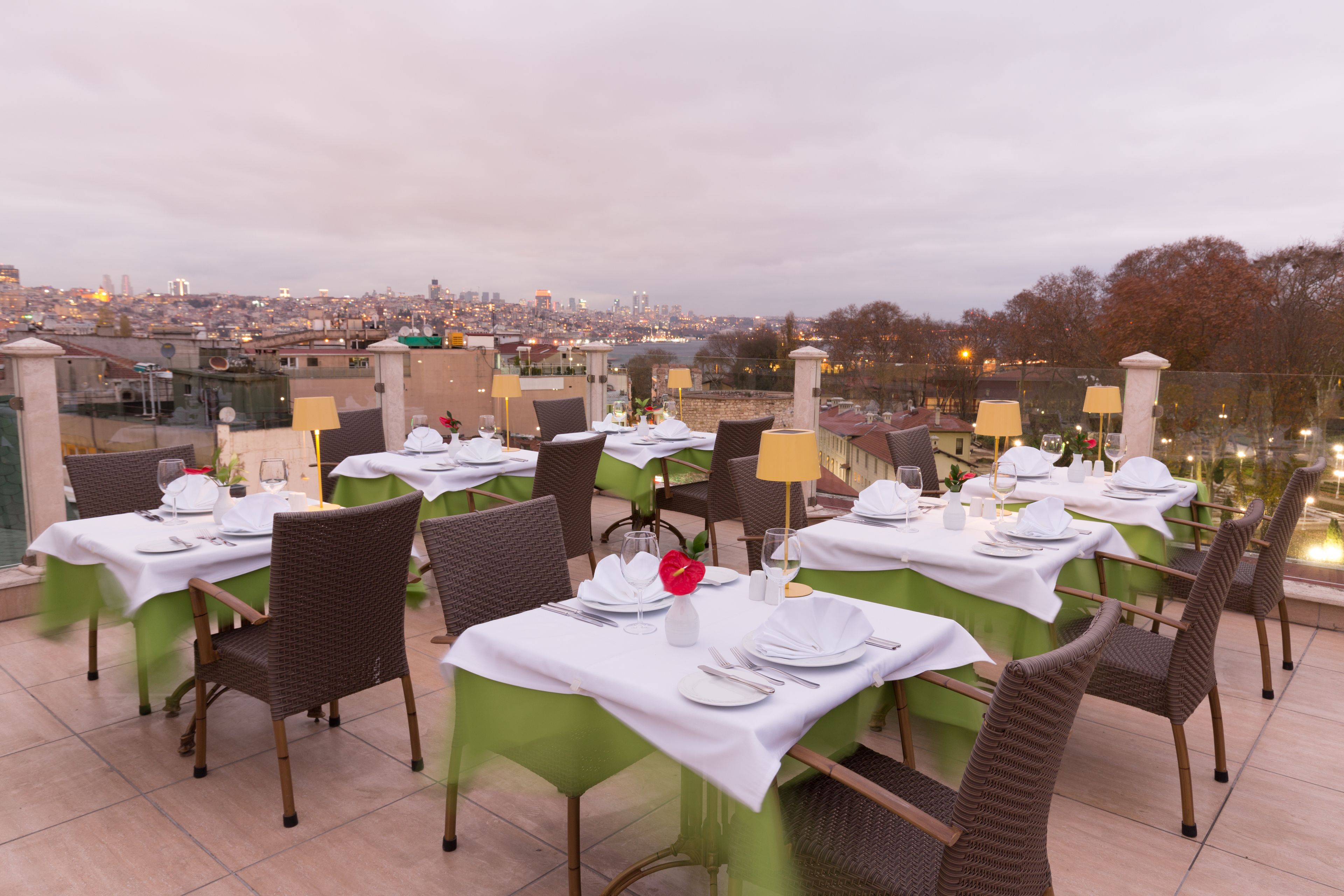 Un área de comedor al aire libre con mesas preparadas para dos, sillas y una vista de la ciudad al fondo.