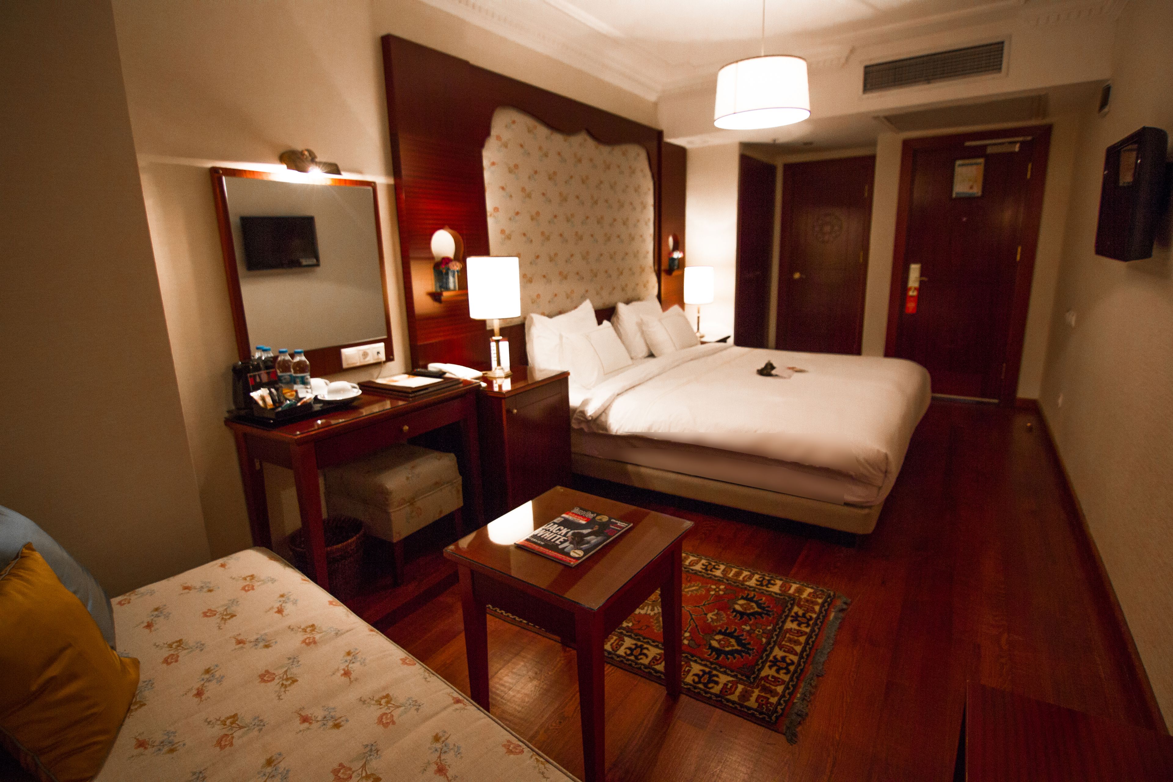 A hotel room with a bed, a desk with a lamp, a chair, a small table, and a television.