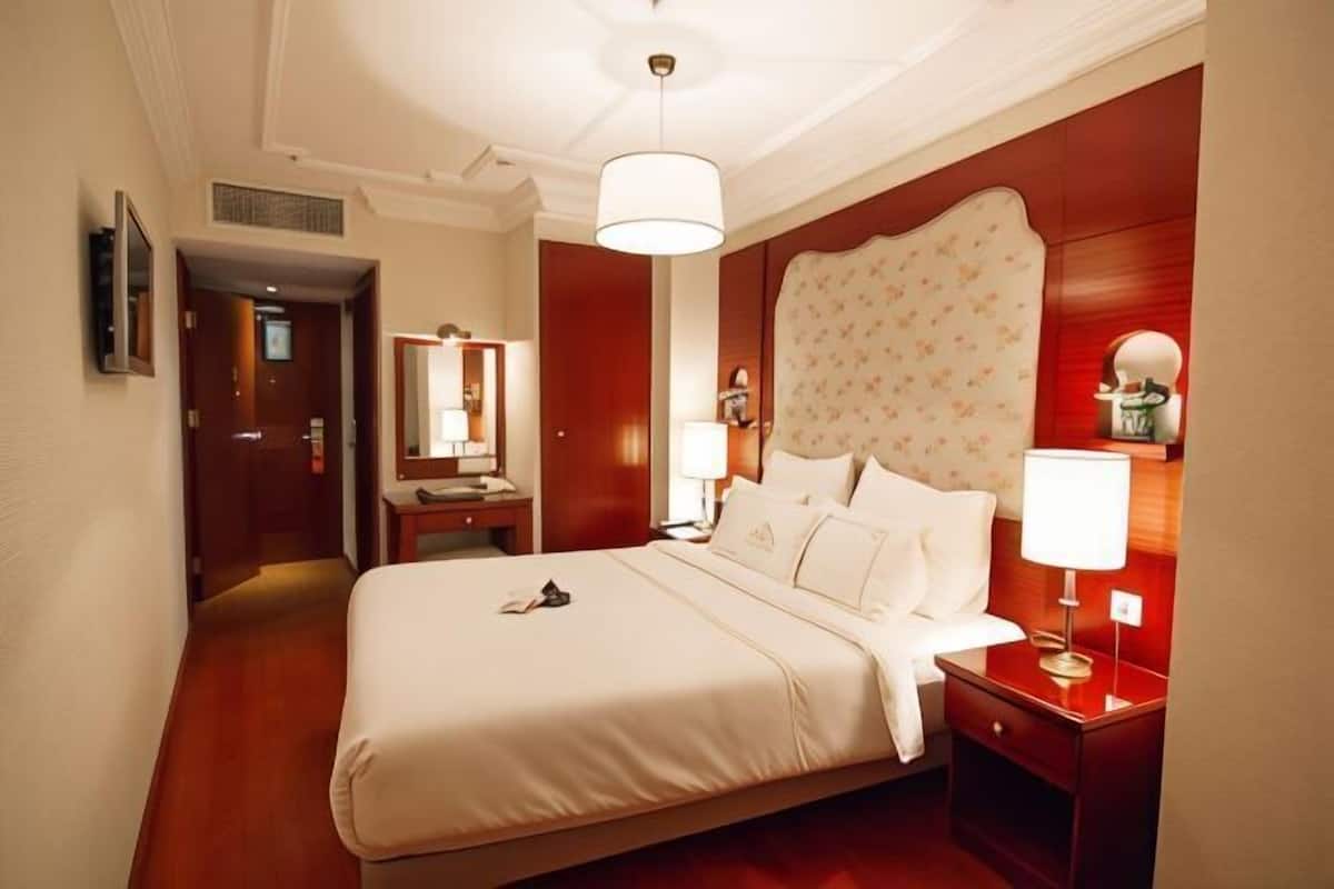 A hotel room with a large bed, bedside tables, a mirror, and a chandelier.