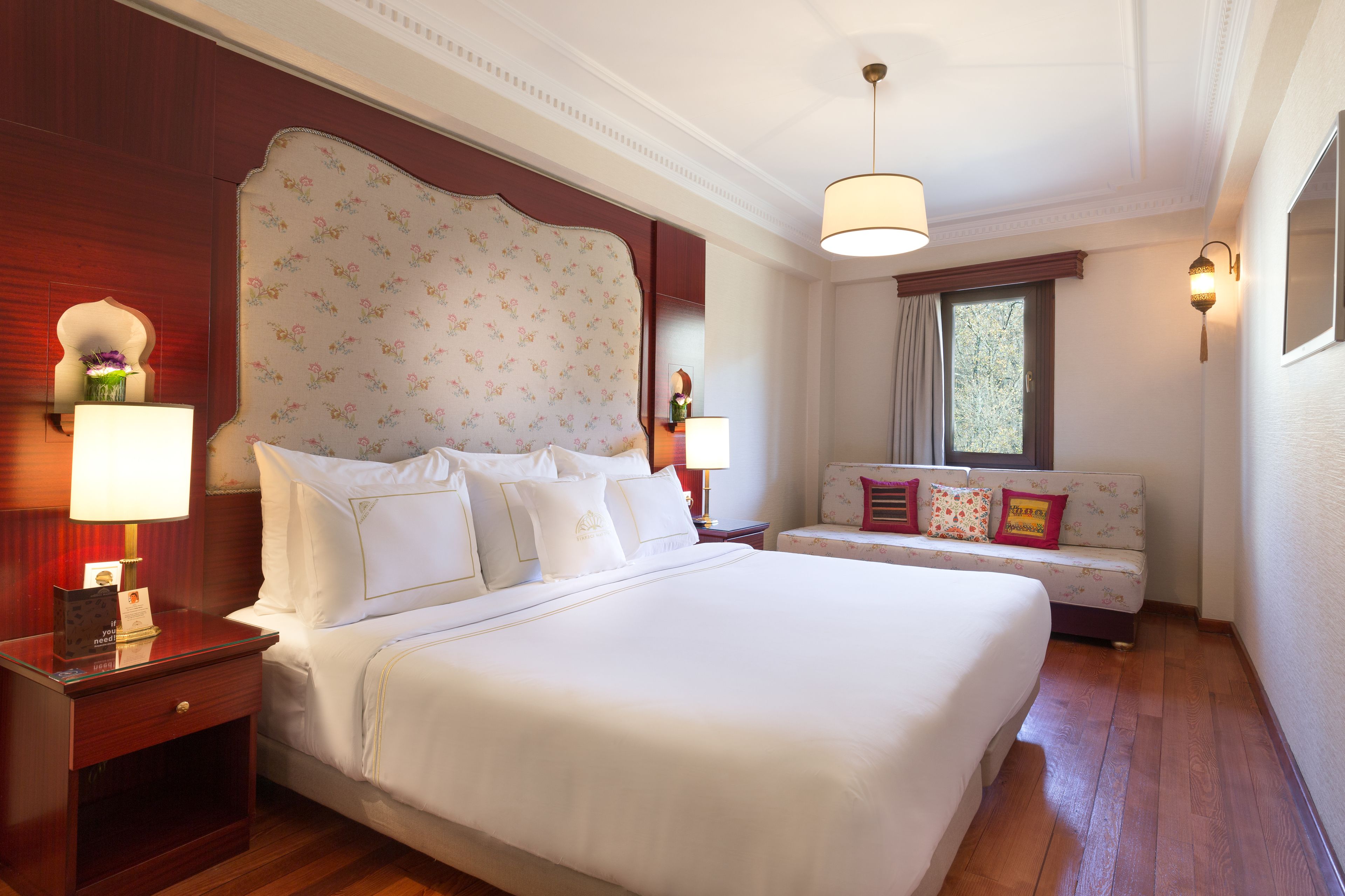 A large bed with white linens, a wooden nightstand, a bedside table with a lamp, a floral-patterned headboard, a window with curtains, and a small sofa with colorful cushions.