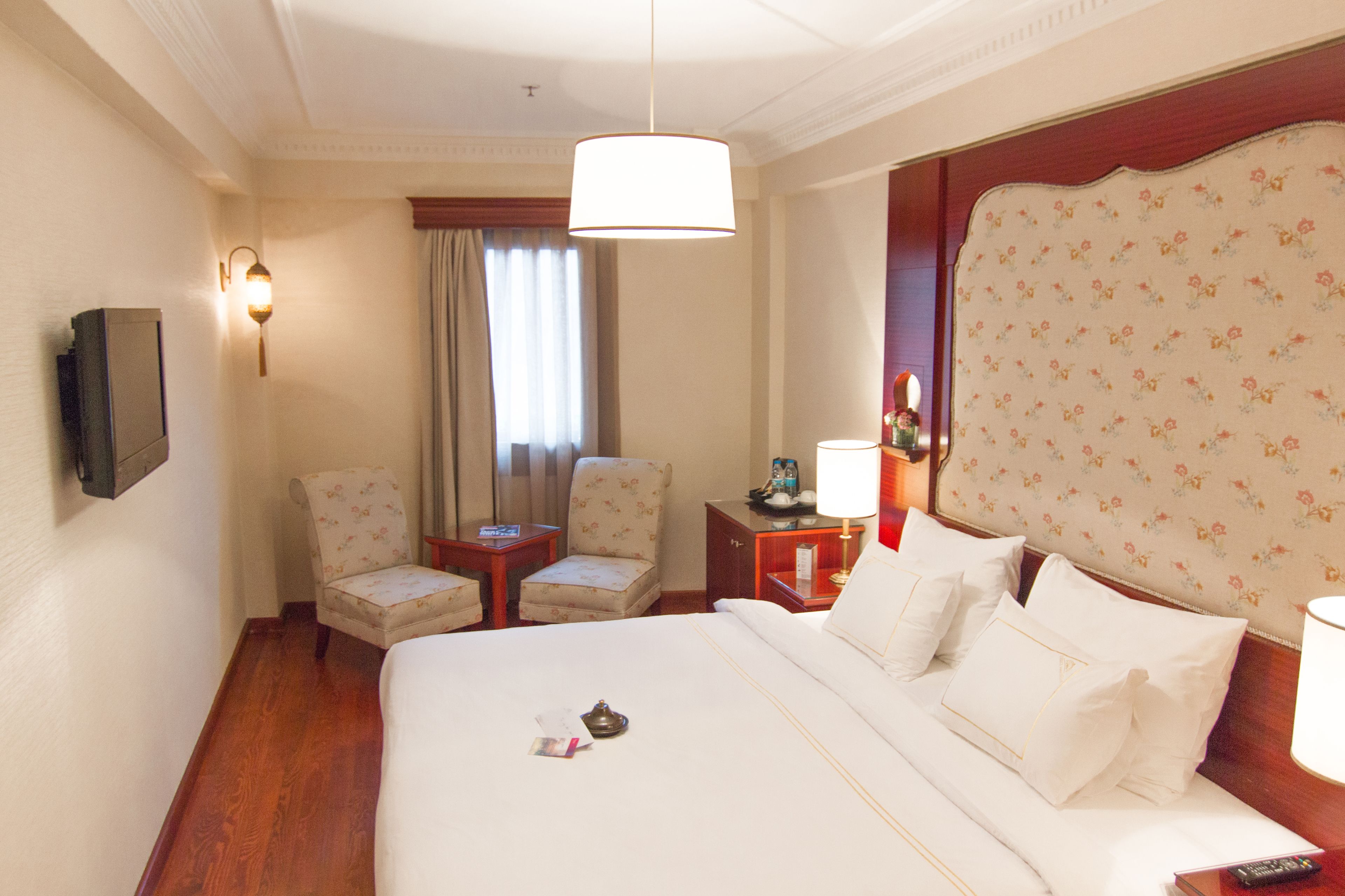 A hotel room with a large bed, two armchairs, a small table, a nightstand with a lamp, and a television mounted on the wall.