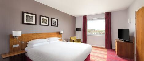 Una habitación de hotel con una cama grande, televisión, escritorio con lámpara, una silla y ventana con cortinas.