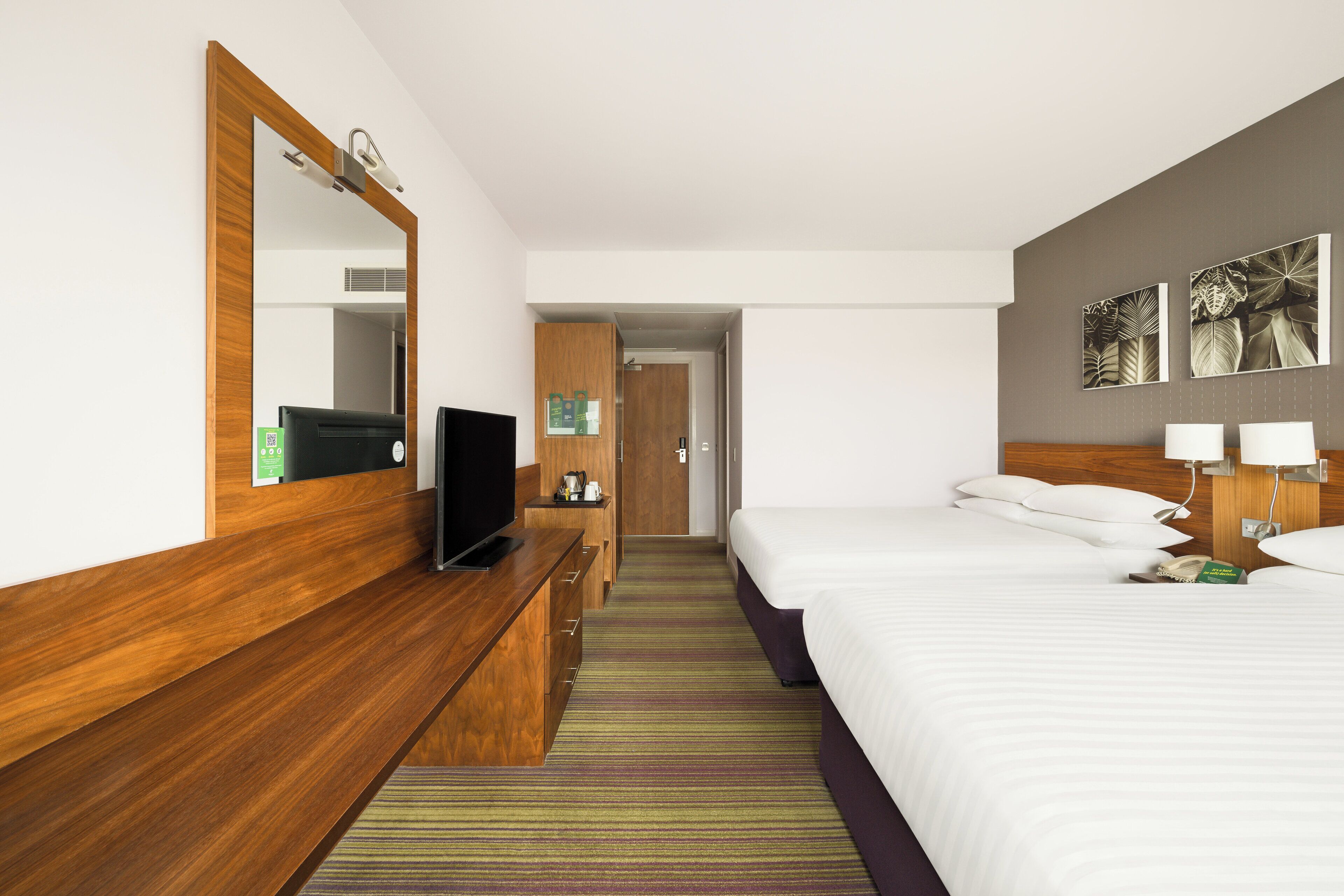 A hotel room with a wooden dresser, a flat-screen TV, a bed with white linens, and a striped carpet.
