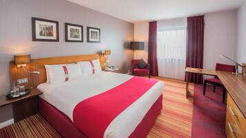 A hotel room with a large bed, a desk, a chair, a nightstand, a lamp, and a window with curtains.