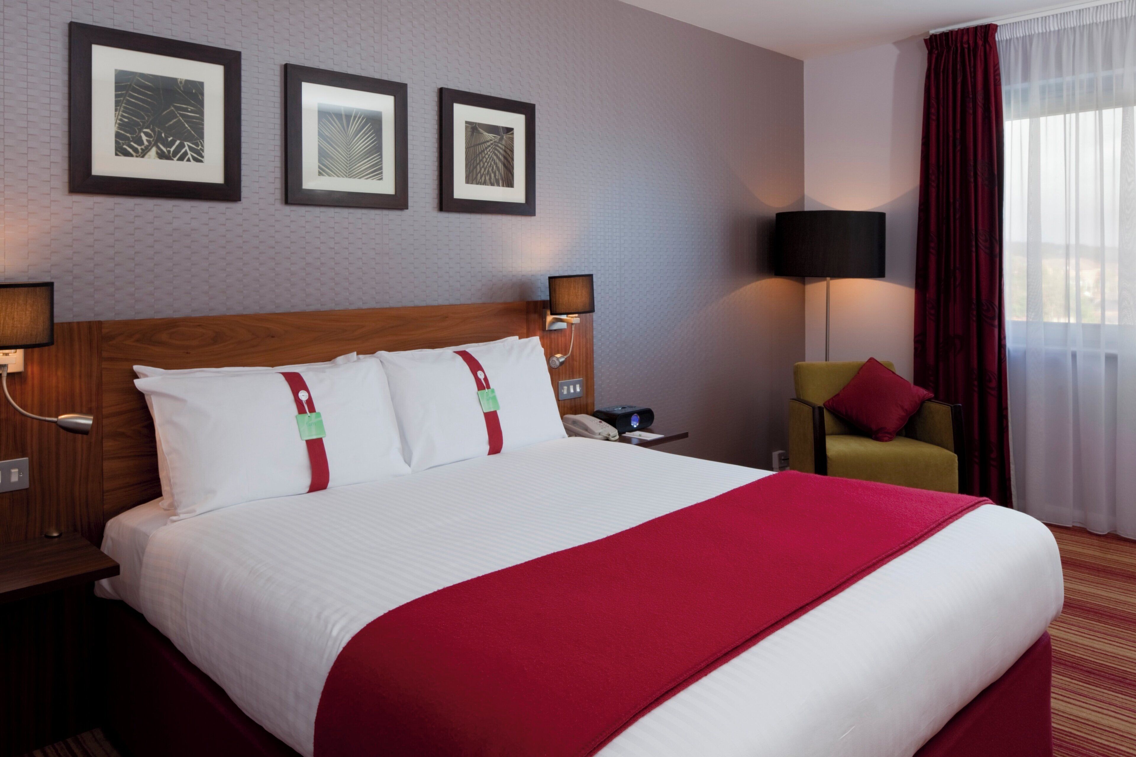 A hotel room with a large bed, a nightstand with a lamp, a chair, and three framed pictures on the wall.