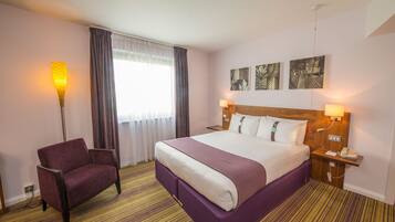 A hotel room with a large bed, a chair, a nightstand with a phone, and artwork on the wall.