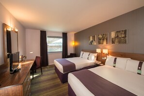 A hotel room with two beds, a desk with a computer, a chair, and artwork on the wall.