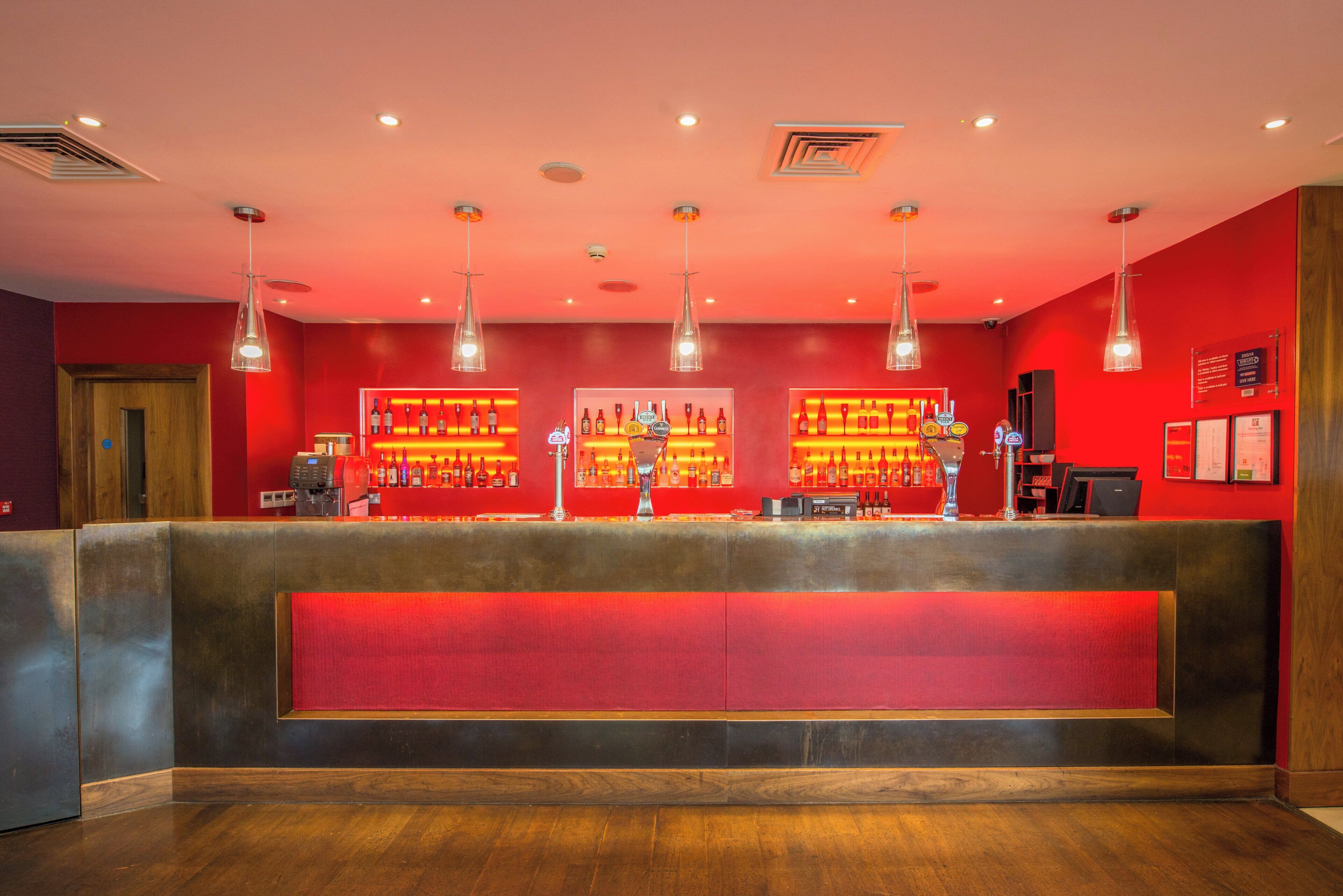 Eine Bar mit roter Wand, mehreren Flaschenregalen und hängenden Pendelleuchten.