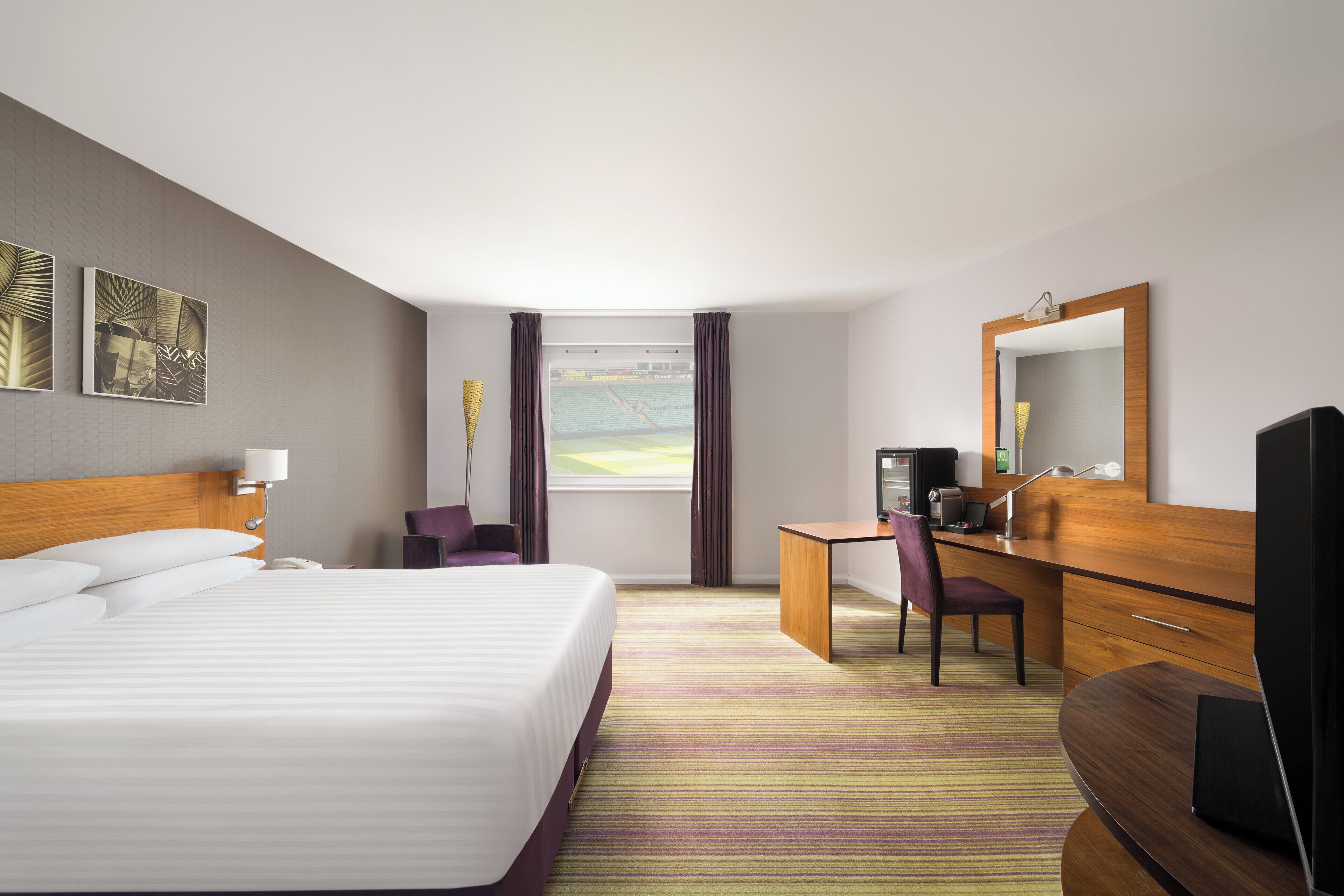 A hotel room with a large bed, a desk with a computer, a chair, a TV, and a window with curtains.