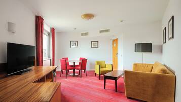 A modern hotel room with a flat-screen TV, a wooden desk, a red carpeted floor, a yellow armchair, a mustard-colored sofa, and a dining area with red chairs.