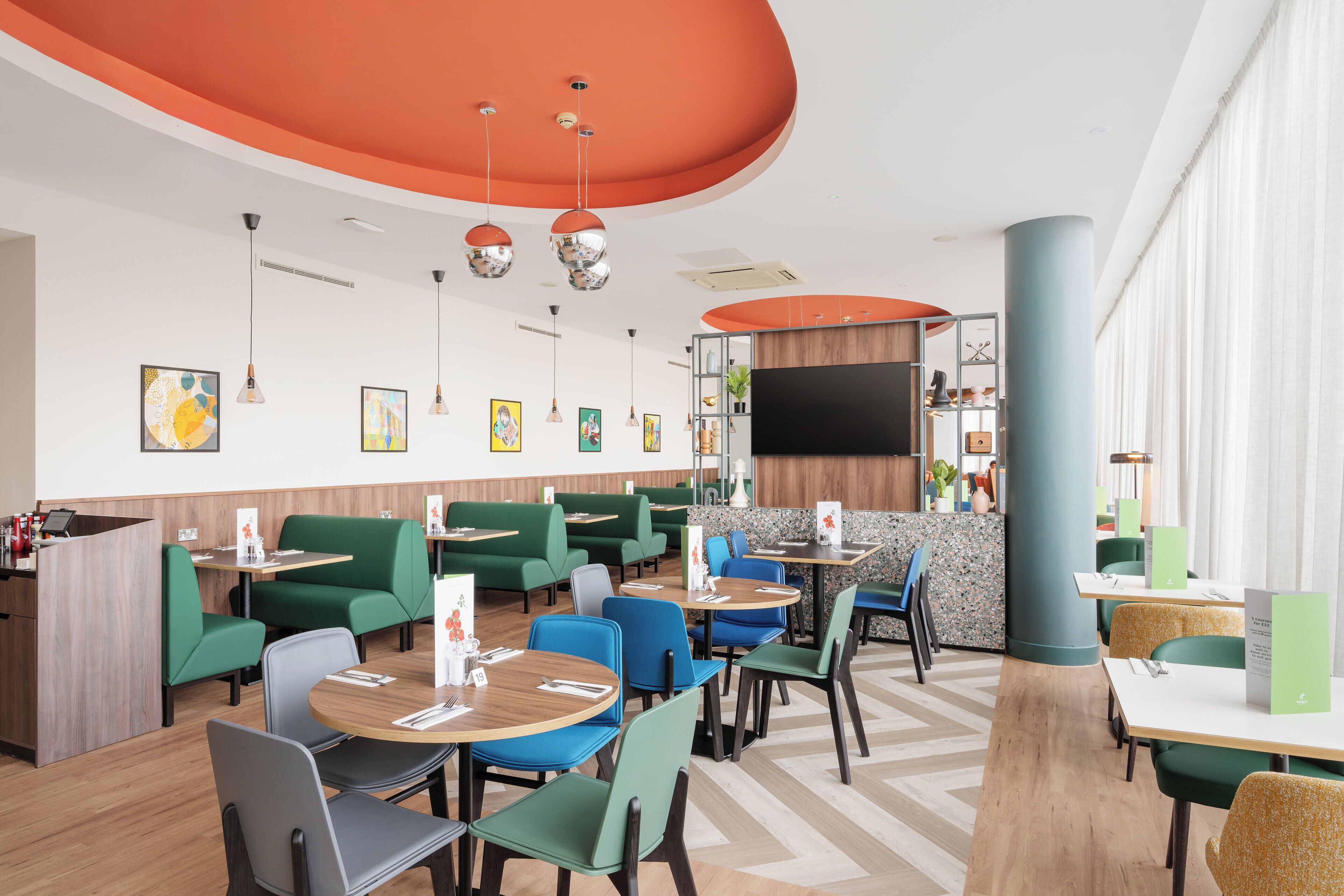 A modern restaurant with green booths, round tables, and a large TV screen.