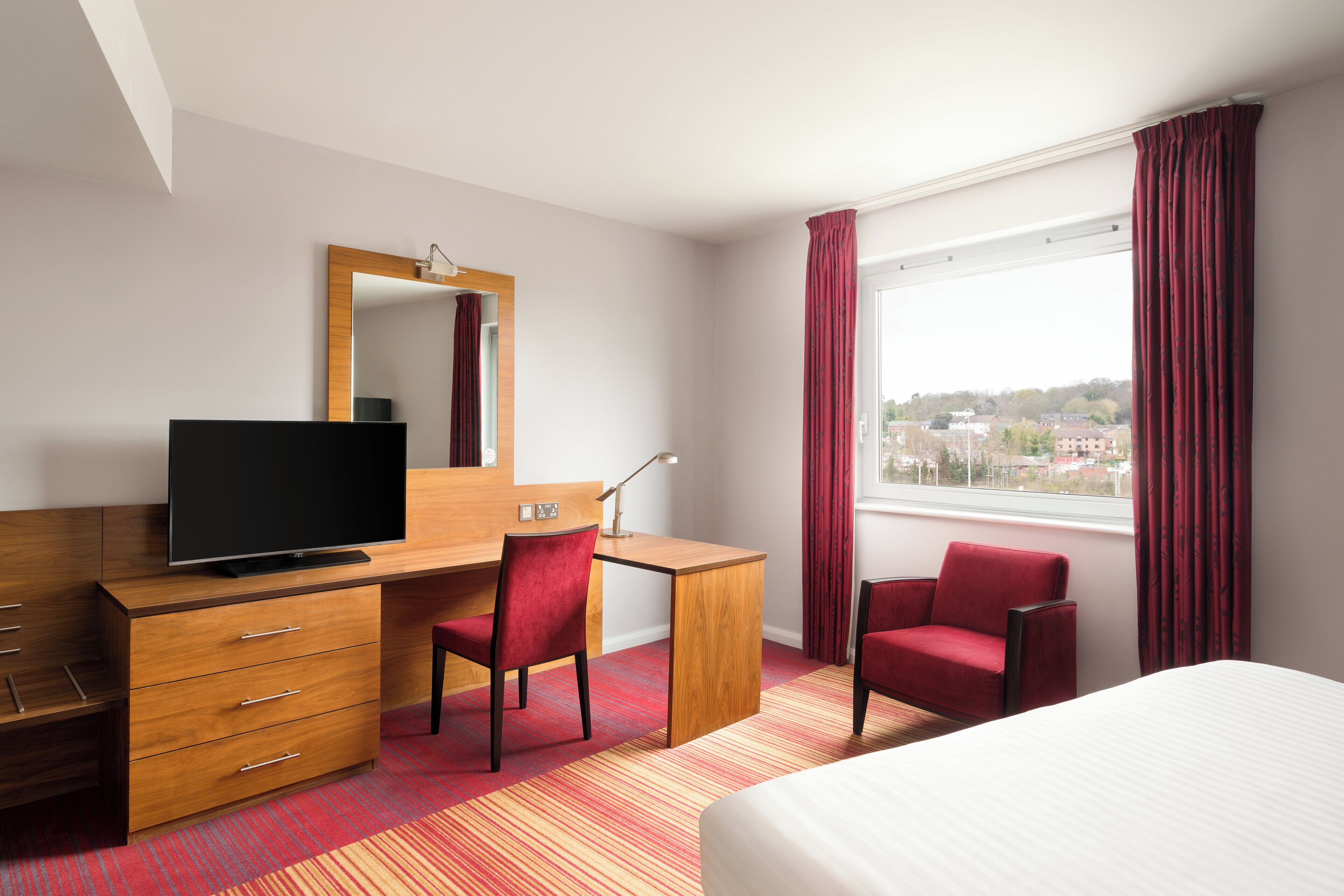 Habitación de hotel con cama, escritorio con televisor, silla, cómoda y un amplio ventanal con cortinas.
