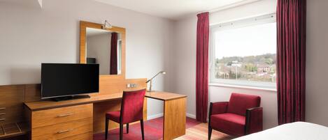 Ein Hotelzimmer mit einem Bett, einem Schreibtisch mit Fernseher, einem Stuhl, einem Schminktisch und einem großen Fenster mit Vorhängen.