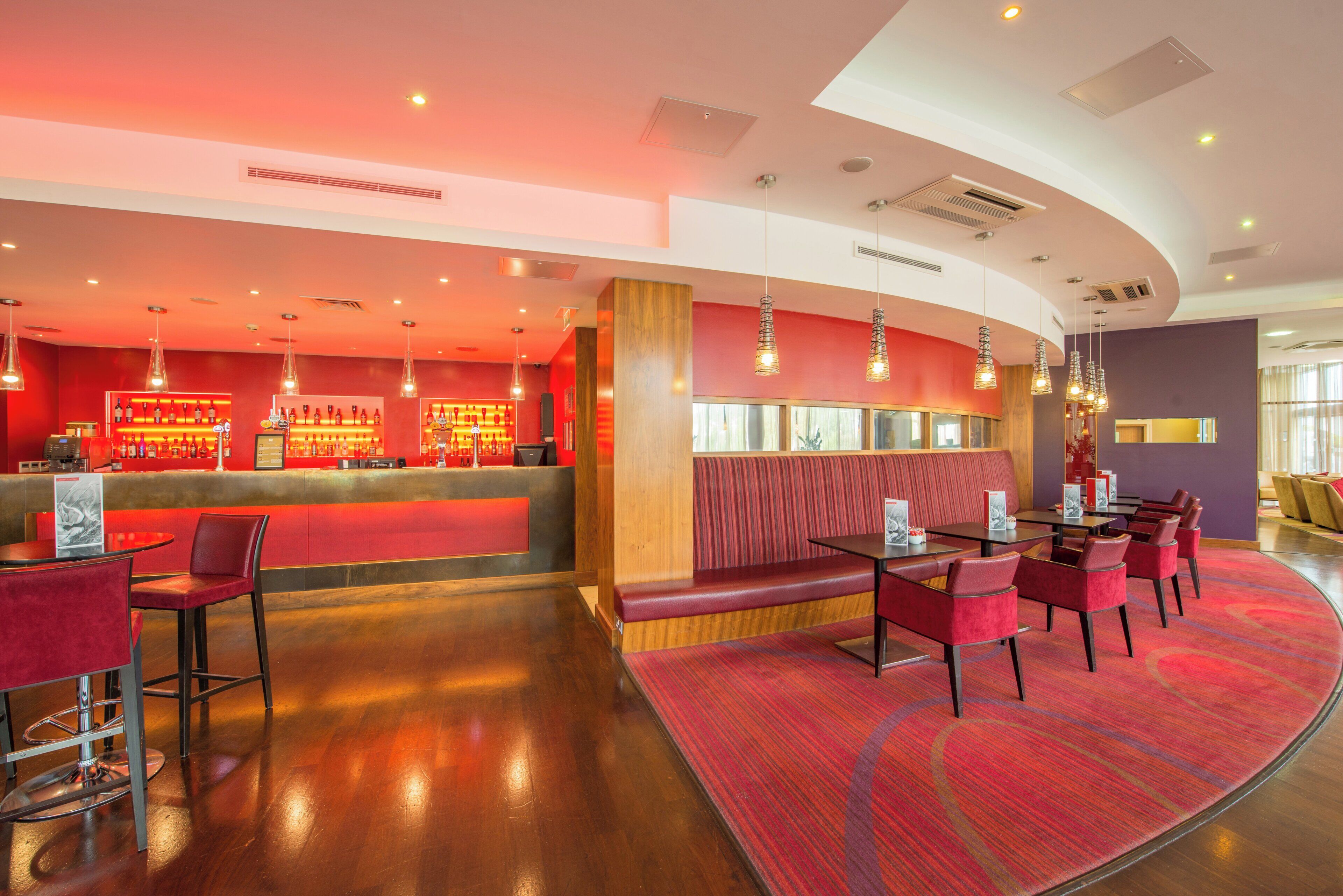 A modern restaurant interior with a curved layout, red seating, a bar area, and pendant lighting.