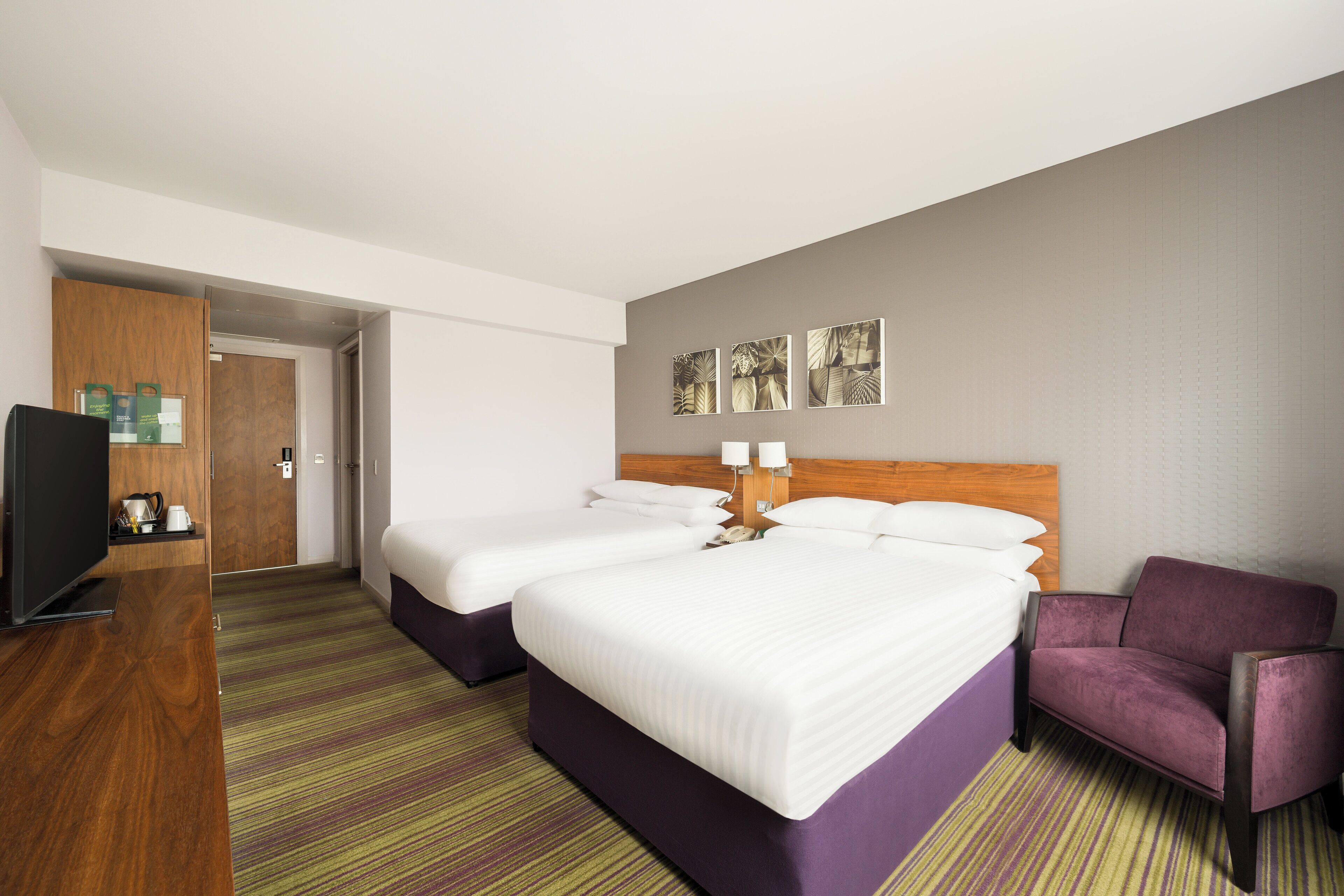 A hotel room with two beds, a TV, a coffee machine, and a purple armchair.