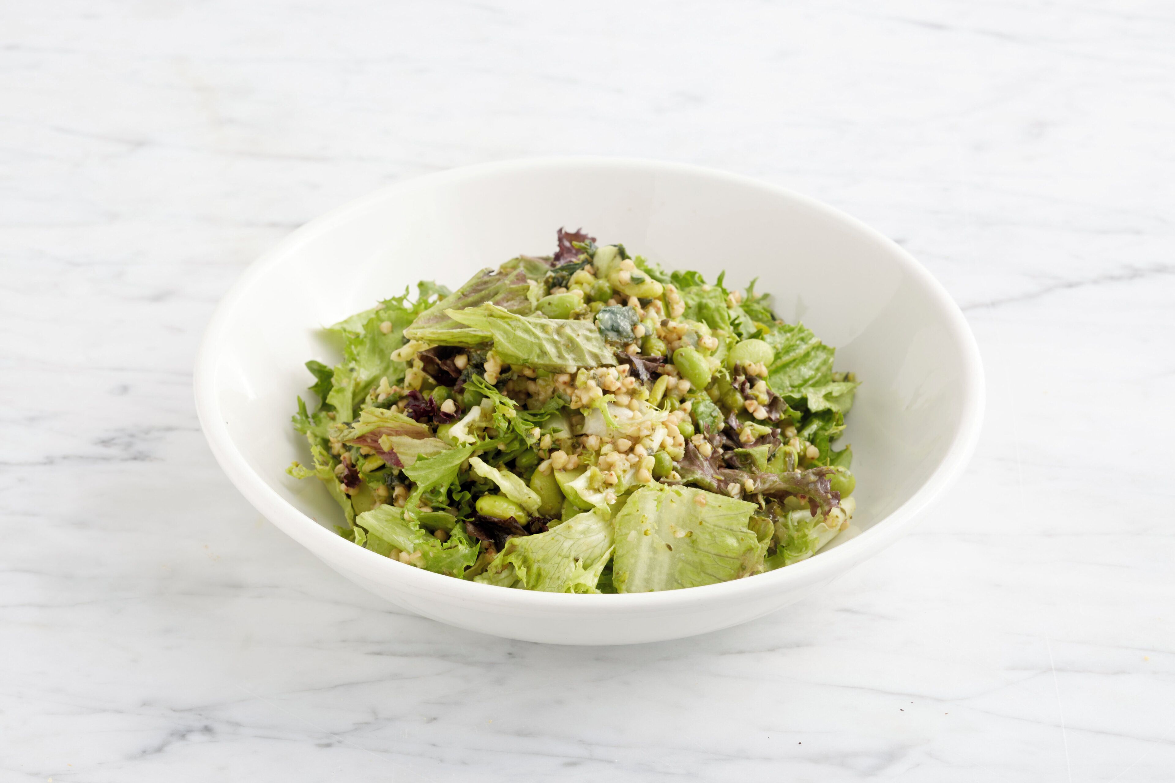 A white bowl containing a salad with various greens and grains on a marble surface.