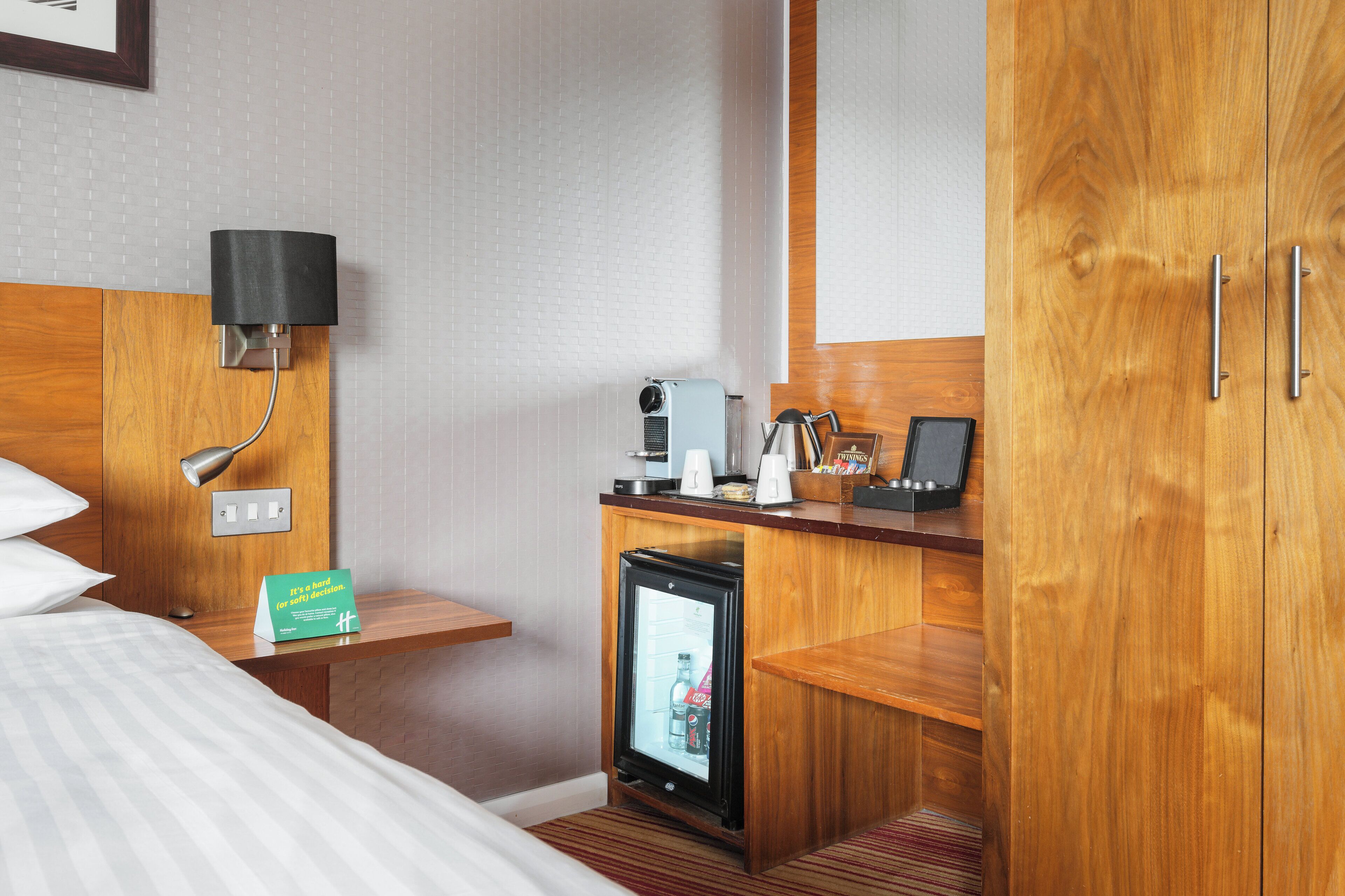 A hotel room with a bed, a wooden nightstand, a minibar, a desk with a lamp, and a wardrobe.