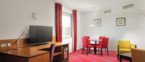 A hotel room with a wooden desk, a flat-screen TV, a red carpeted floor, a dining table with red chairs, and a yellow armchair.