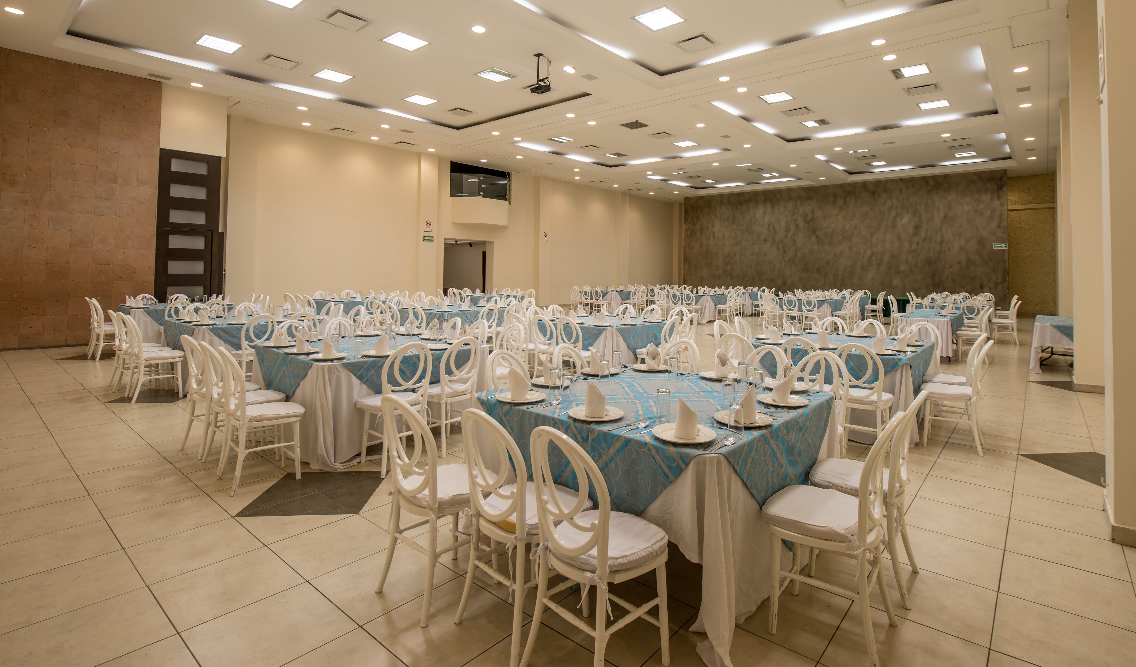 Un amplio salón de conferencias con varias mesas dispuestas para un evento formal, equipado con sillas blancas y mesas cubiertas con manteles azules.