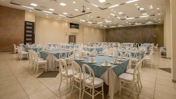 Un amplio salón de conferencias con varias mesas dispuestas para un evento formal, equipado con sillas blancas y mesas cubiertas con manteles azules.