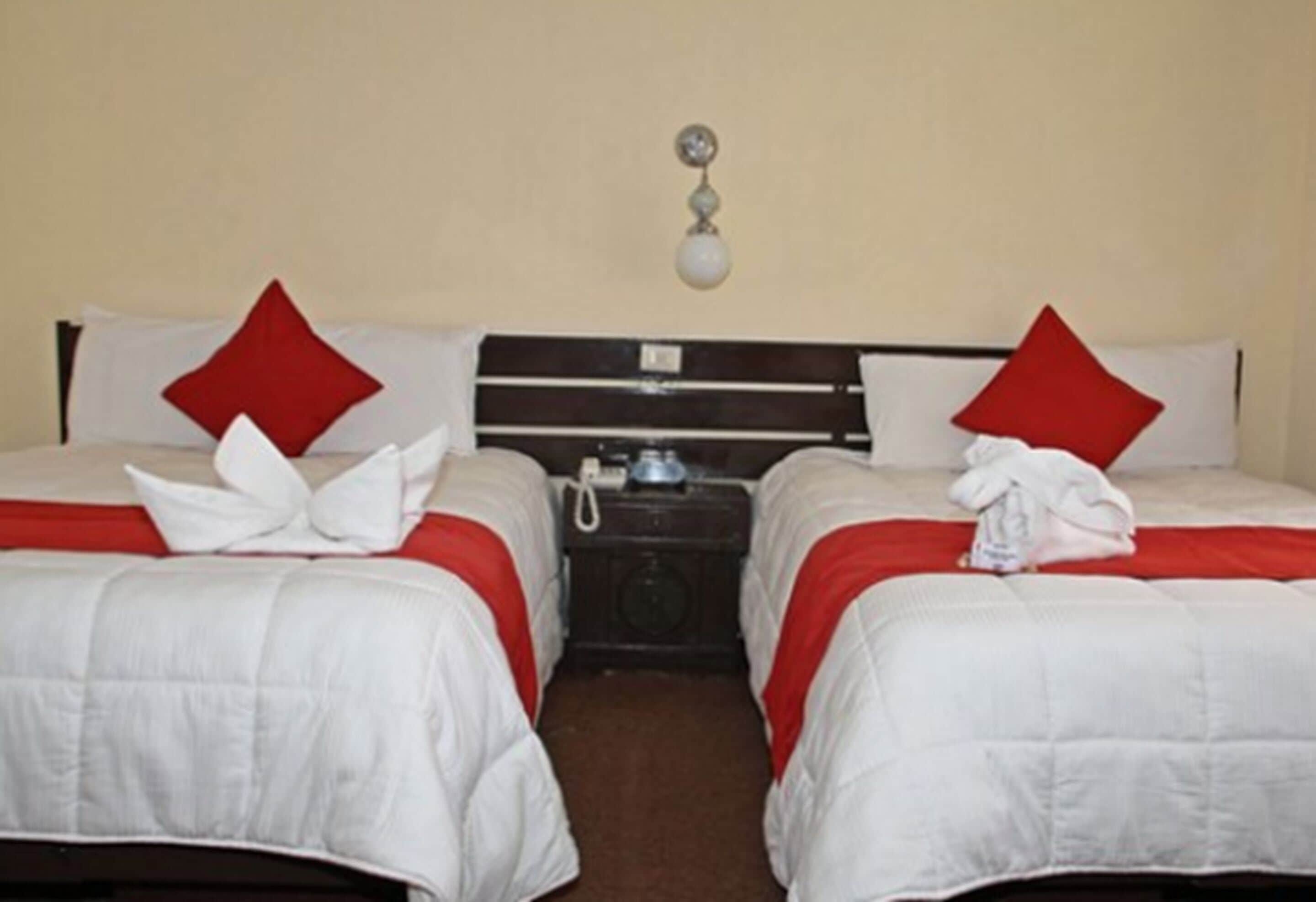 A hotel room with two beds, each with a red pillow and white bedding. There is a nightstand between the beds with a lamp and a phone.