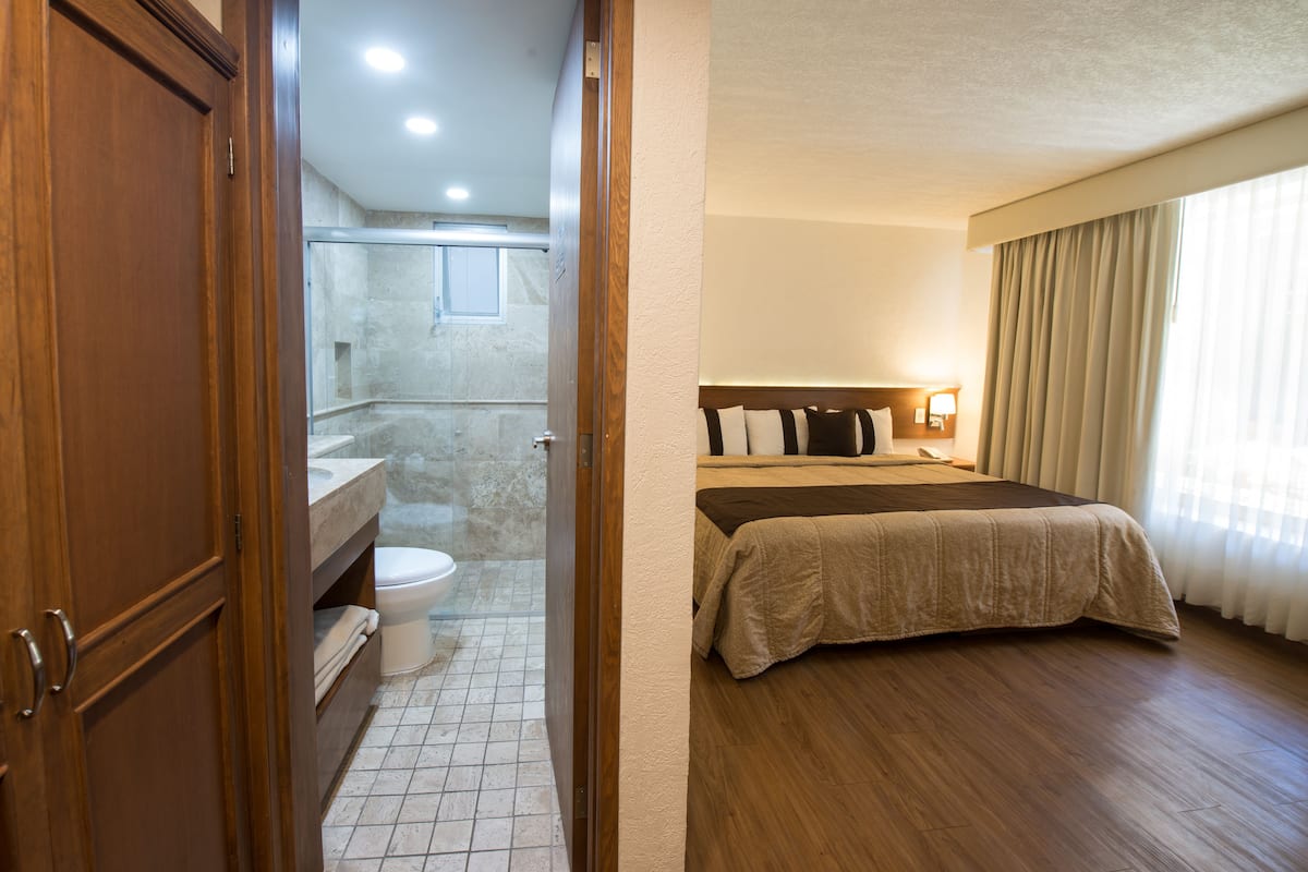 Habitación de hotel con suelo de madera, cama con colcha color beige, mesita de noche con lámpara y baño con inodoro y lavamanos.