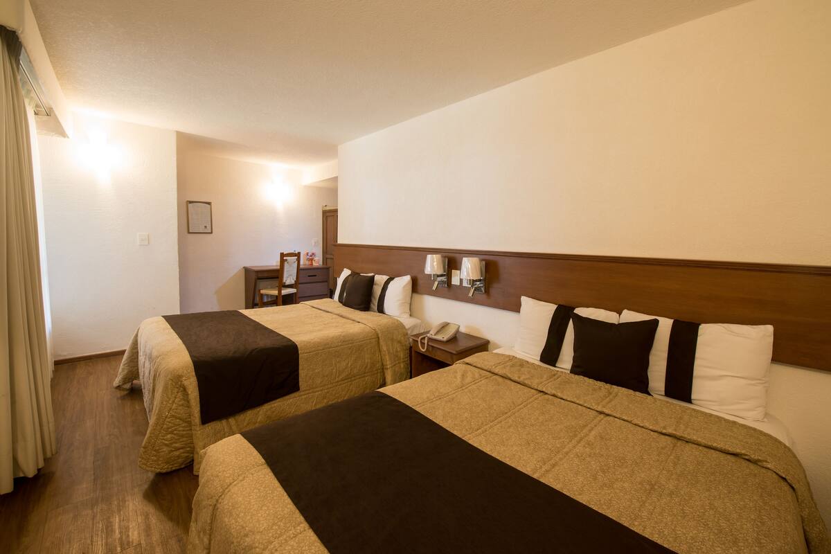 Habitación de hotel con dos camas, cabecero de madera, lámparas de noche, teléfono y una pequeña mesa con una silla.