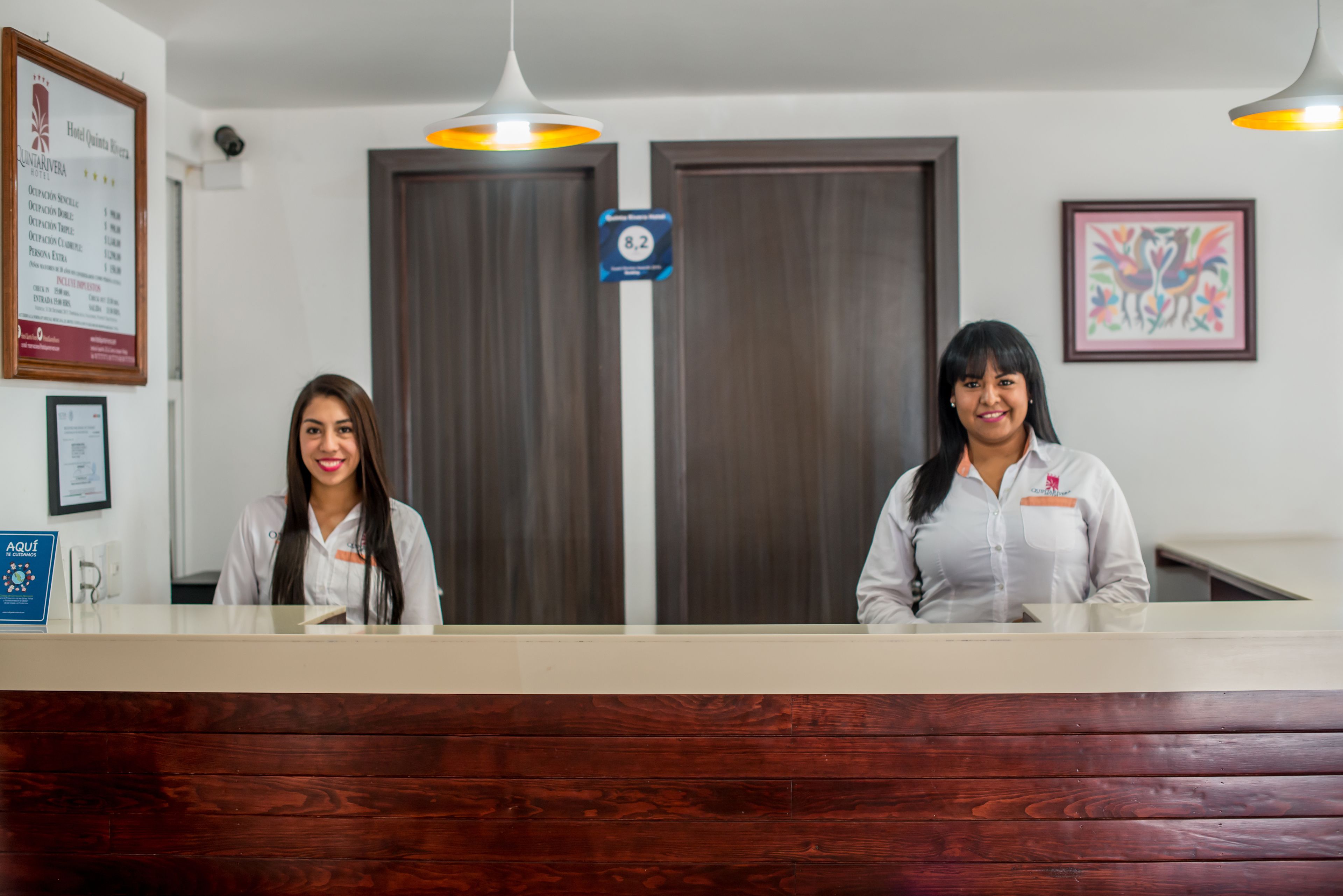 Dos recepcionistas en el mostrador de recepción de un hotel, con un mostrador de madera, diplomas enmarcados y un cartel que indica la habitación 8.2.