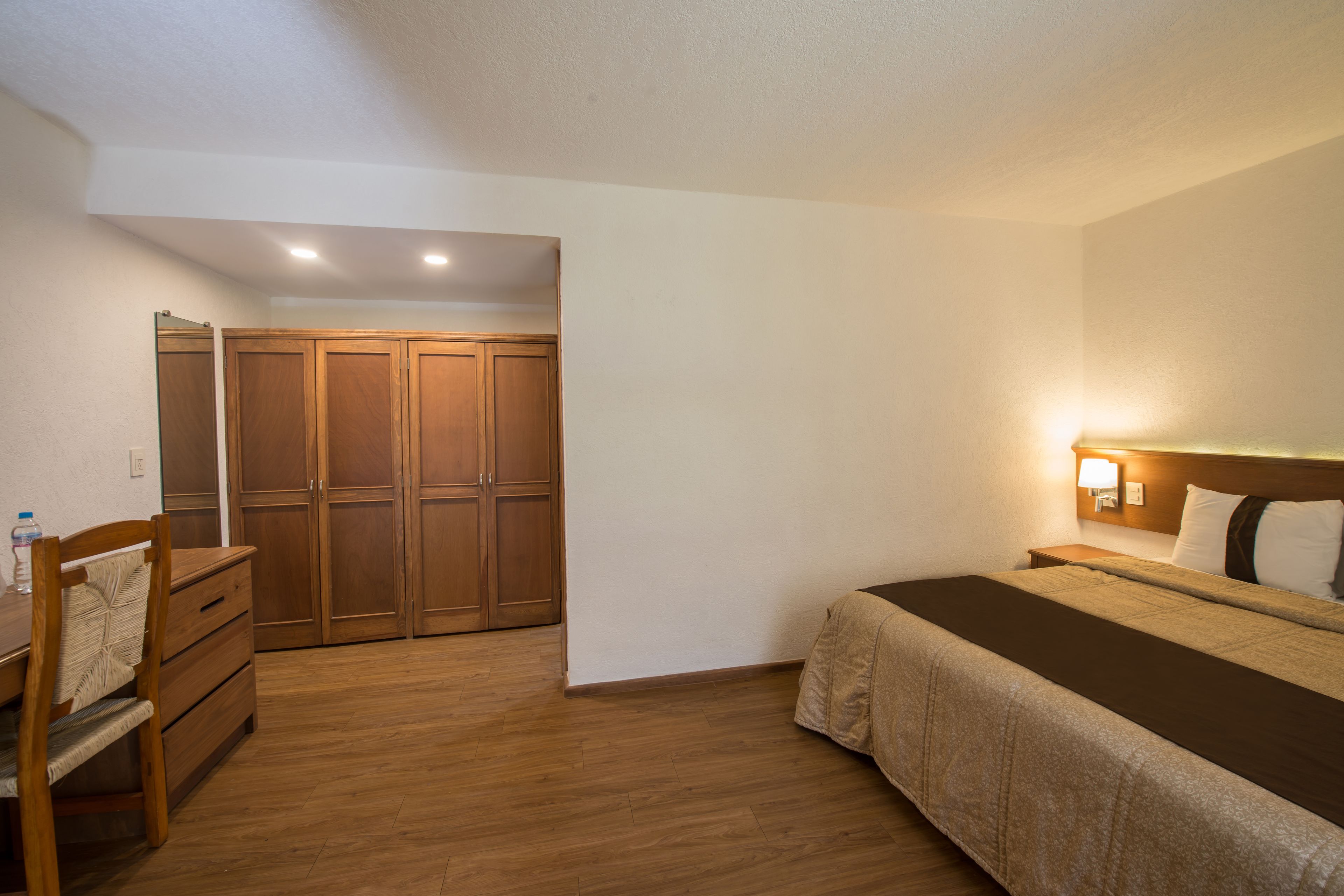 Een hotelkamer met een houten vloer, een bed met een bruine plaid, een houten bureau met stoel en een garderobe met houten deuren.
